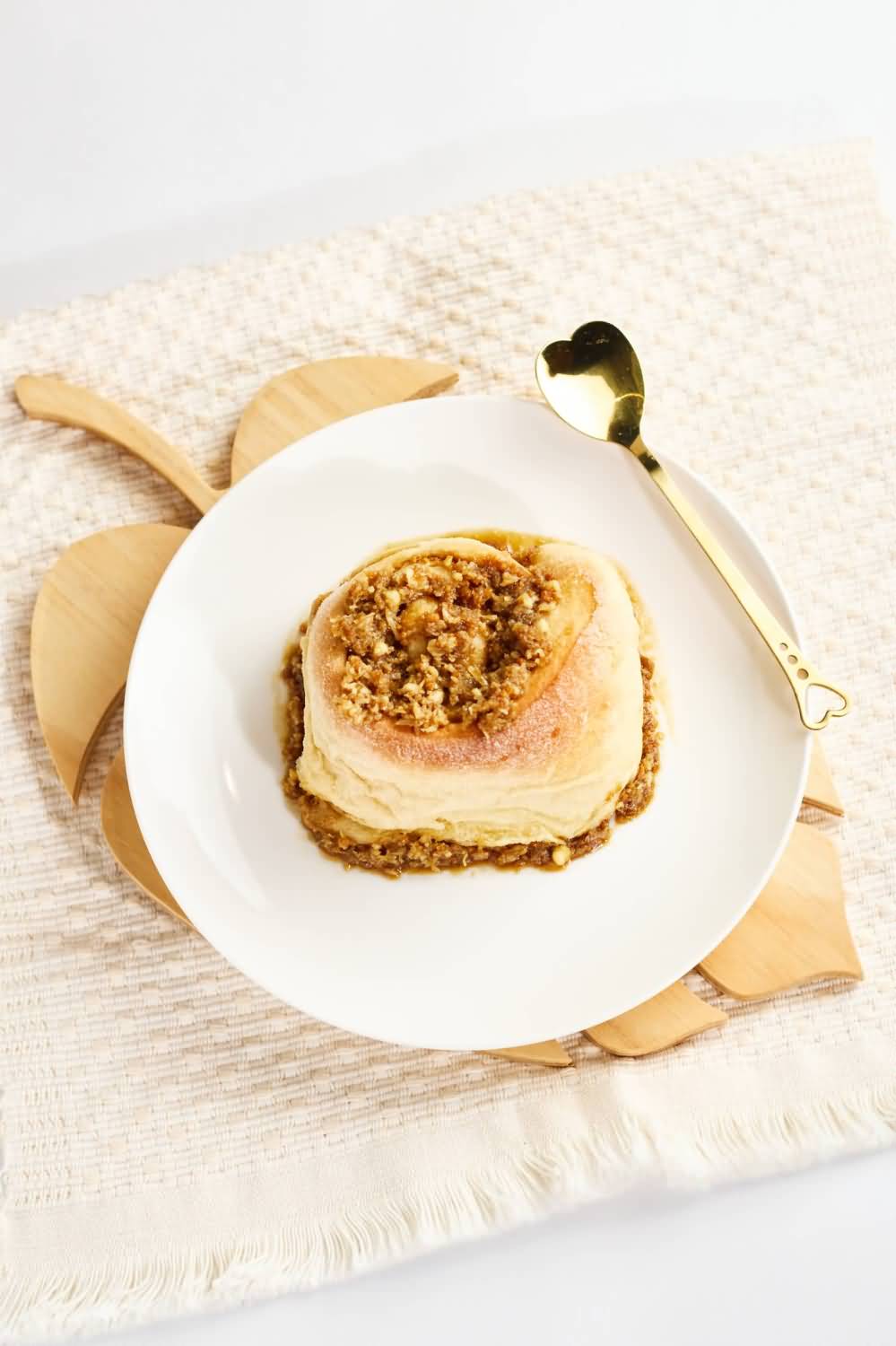 Un cinnamon roll esponjoso con un relleno de canela y nueces caramelizadas, hecho en la ciudad de Medellín, servido en un plato blanco sobre un mantel texturizado color crema, acompañado de una cuchara dorada decorativa con forma de corazón en el mango.